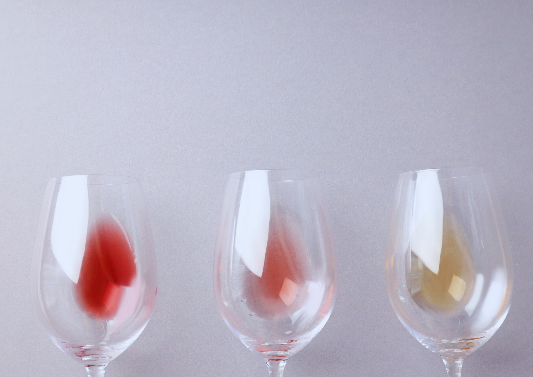 Drei leere Weingläser mit Farbspuren von Rot-, Rosé- und Weißwein auf grauem Hintergrund.