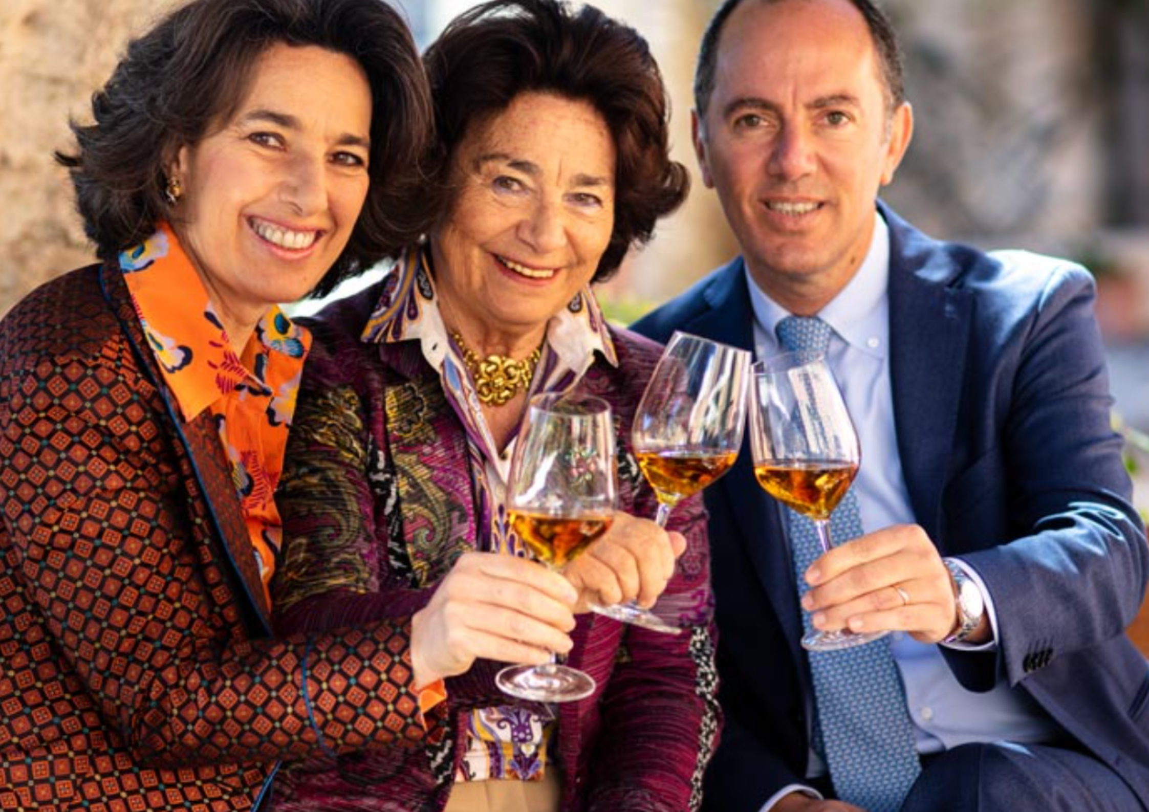 Drei elegant gekleidete Personen stoßen draußen fröhlich mit Weißweingläsern an.