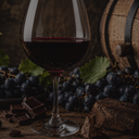 „Glas Rotwein mit dunklen Trauben, Schokolade und Holzfass im Hintergrund“
